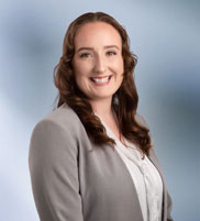 woman, Destiny Hughes, wearing a light gray blazer and white blouse, standing against a softly blurred blue and white background, smiling confidently at the camera