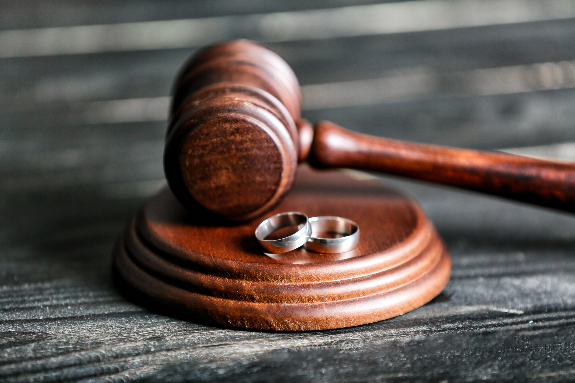 Wood Gavel and pounding block with two silver wedding rings on block and resting on black wooden table.