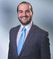 Man, Patrick Quinn, with short brown hair wearing a dark gray blazer, smiling against a blurred white and blue background.