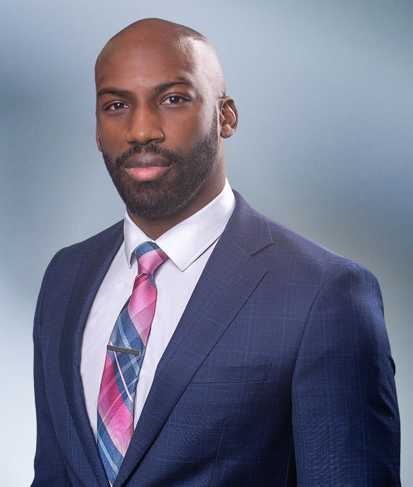 A bald, black man, Xavier Prather, wearing a dark gray blazer, smiling against a blurred white and blue background.
