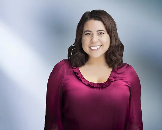 A woman, Jacquelyn Dupler, in a deep magenta blouse with ruffled detailing is smiling at the camera, standing against a blurred blue and white background.
