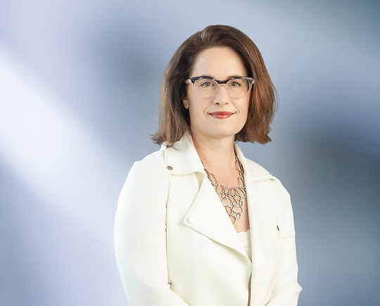 A woman, Andrea Badalucco, in a white jacket, glasses and chain link necklace is smiling at the camera, standing against a blurred blue and white background.