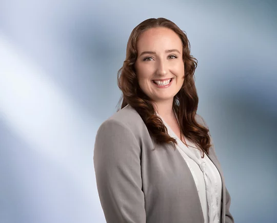 woman, Destiny Hughes, wearing a light gray blazer and white blouse, standing against a softly blurred blue and white background, smiling confidently at the camera