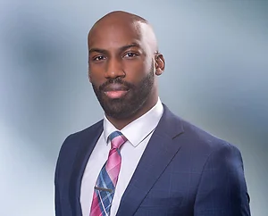 Bald bearded African American man, Xavier Prather, smiles at camera wearing a blue blazer, white undershirt and pink and blue striped necktie in front of blue gray blurred background.